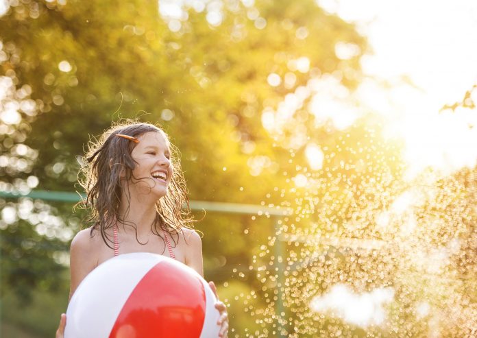 Smiling girl outdoors holding a large red and white beach ball in warm sunlight.