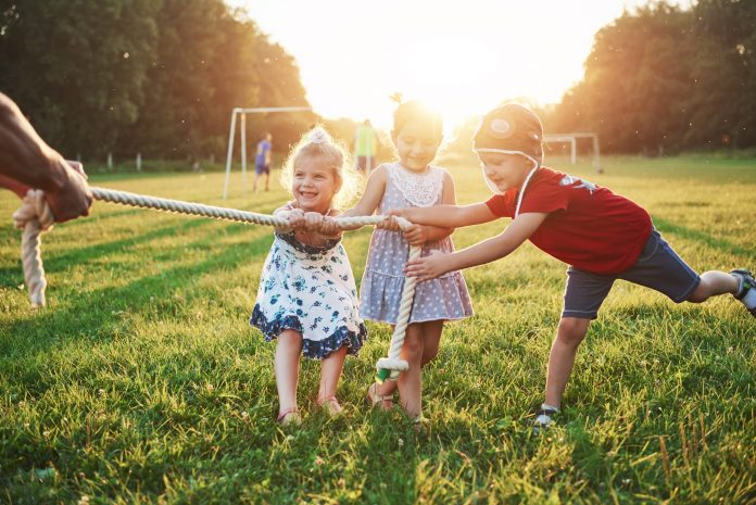 Three children pull a rope together in a grassy field during a sunny outdoor game, smiling faces visible (tug-of-war).